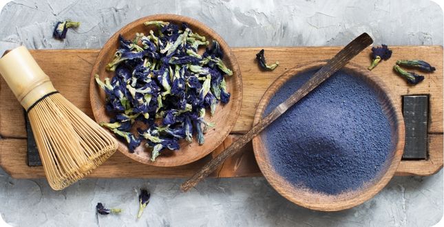 Op een rij liggen een bamboe klopper, een houten schaaltje met gedroogde bloemblaadjes en vervolgens een houten schaaltje met gemalen 'blauwe matcha'.