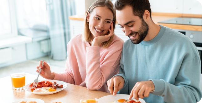 Een lachend stel eet samen een gezond eiwitrijk ontbijt met eieren, groenten en een glas vers sinaasappelsap aan de keukentafel.