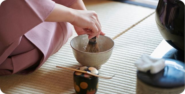 Vrouw die op de grond zit en in een kom japanse matcha opklopt met een bamboe matcha klopper.
